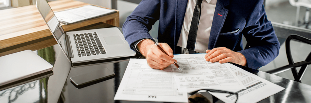 Homem de terno azul escuro e gravata preta, escrevendo em um documento de papel e um notebook aberto em segundo plano
