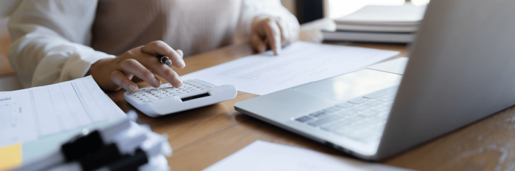 Pessoa realizando cálculos em uma calculadora brnca em frente ao um notebook cinza claro.