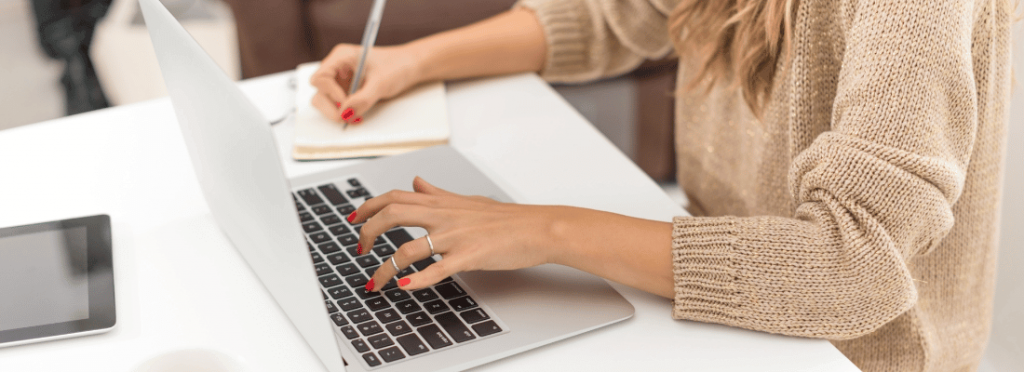 Mulher branca, utilizando um notebook e escrevendo em um caderno ao mesmo tempo.